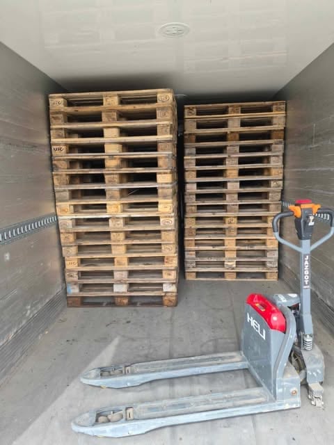Interior of a transport truck cargo area with stacked wooden pallets and a hydraulic hand pallet jack in the foreground