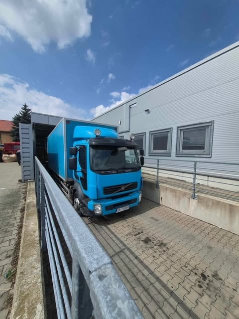 Bright blue Volvo delivery truck parked outside an industrial building with white corrugated walls under a partly cloudy sky