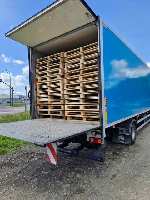 Open blue delivery truck with wooden pallets stacked inside and rear gate lowered, parked outdoors with clear sky