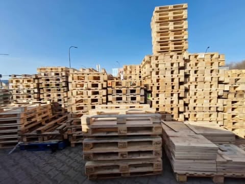 Stacked wooden pallets in warehouse yard with brick apartment buildings in background under clear sky